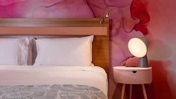 Una cama bien hecha con sábanas blancas y una manta decorativa, una mesita de noche con lámpara, y un mural abstracto y llamativo en la pared.
