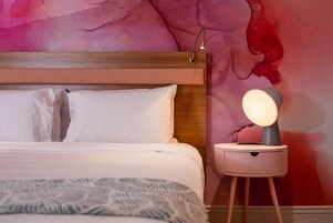 Una cama bien hecha con sábanas blancas y una manta decorativa, una mesita de noche con lámpara, y un mural abstracto y llamativo en la pared.