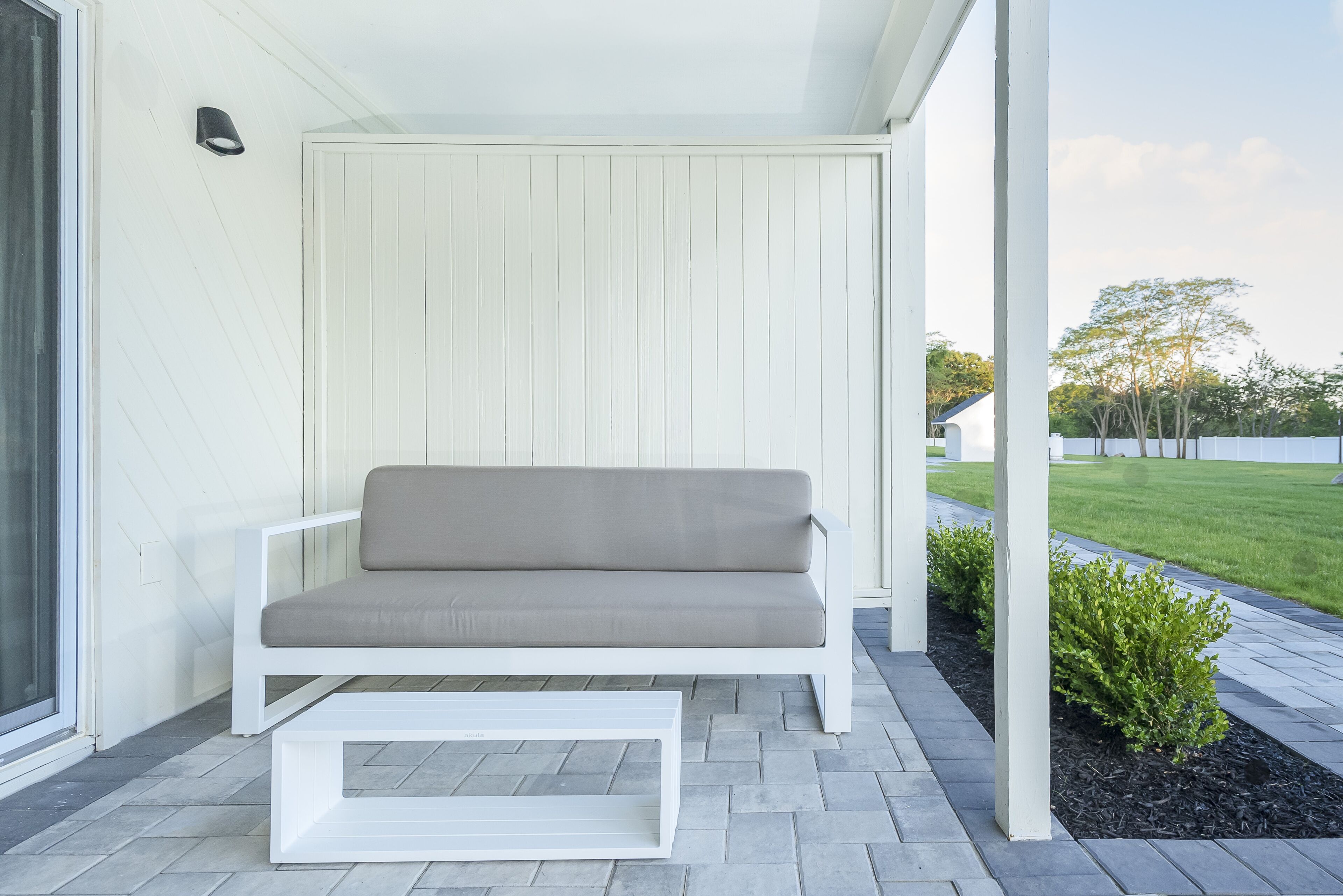 A white outdoor seating area with a bench and a small table, surrounded by a white wall and a paved patio.