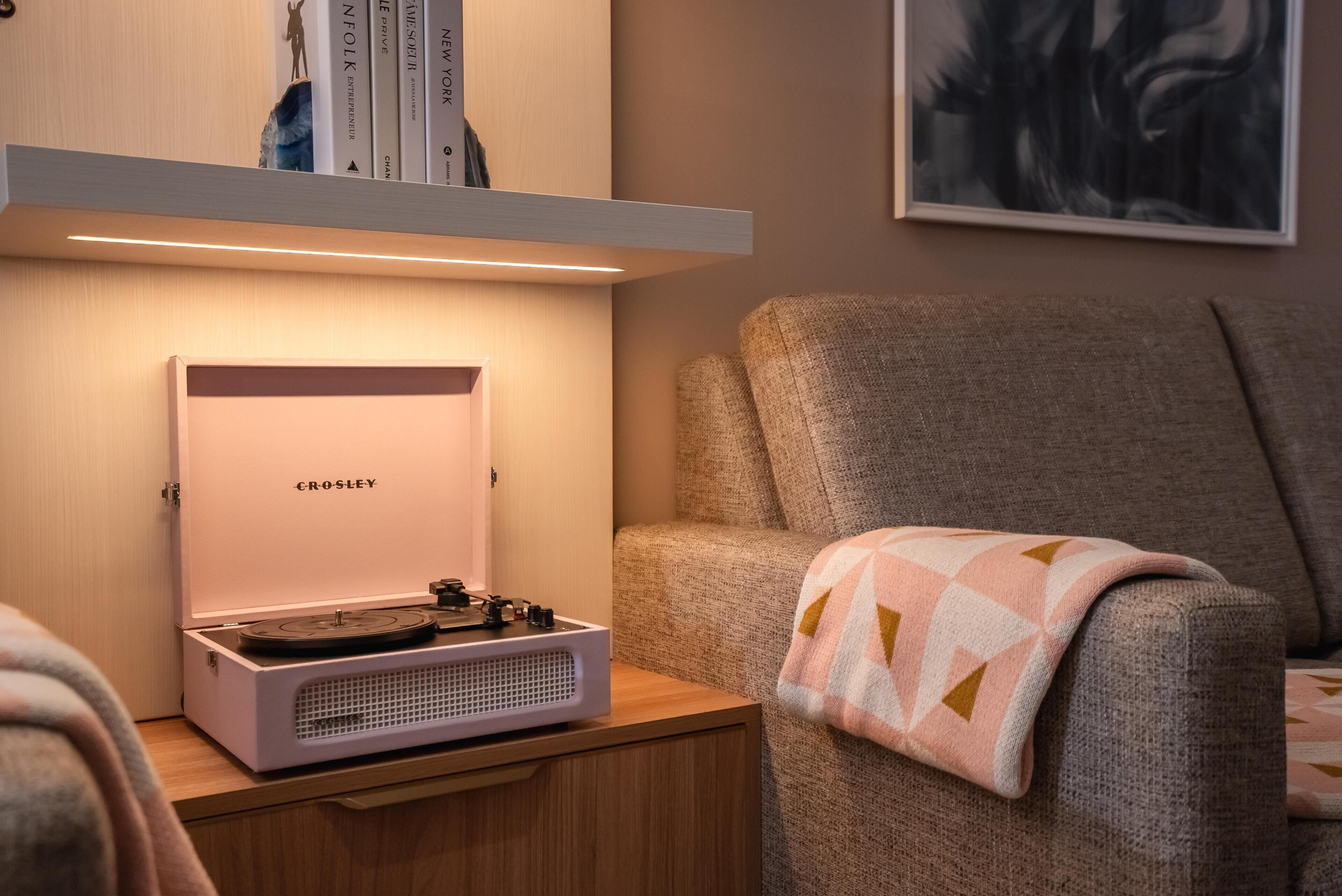 Una habitación acogedora con un reproductor de discos Crosley portátil sobre una mesita de madera, un sillón cómodo con una manta estampada y una estantería con libros y un cuadro enmarcado.