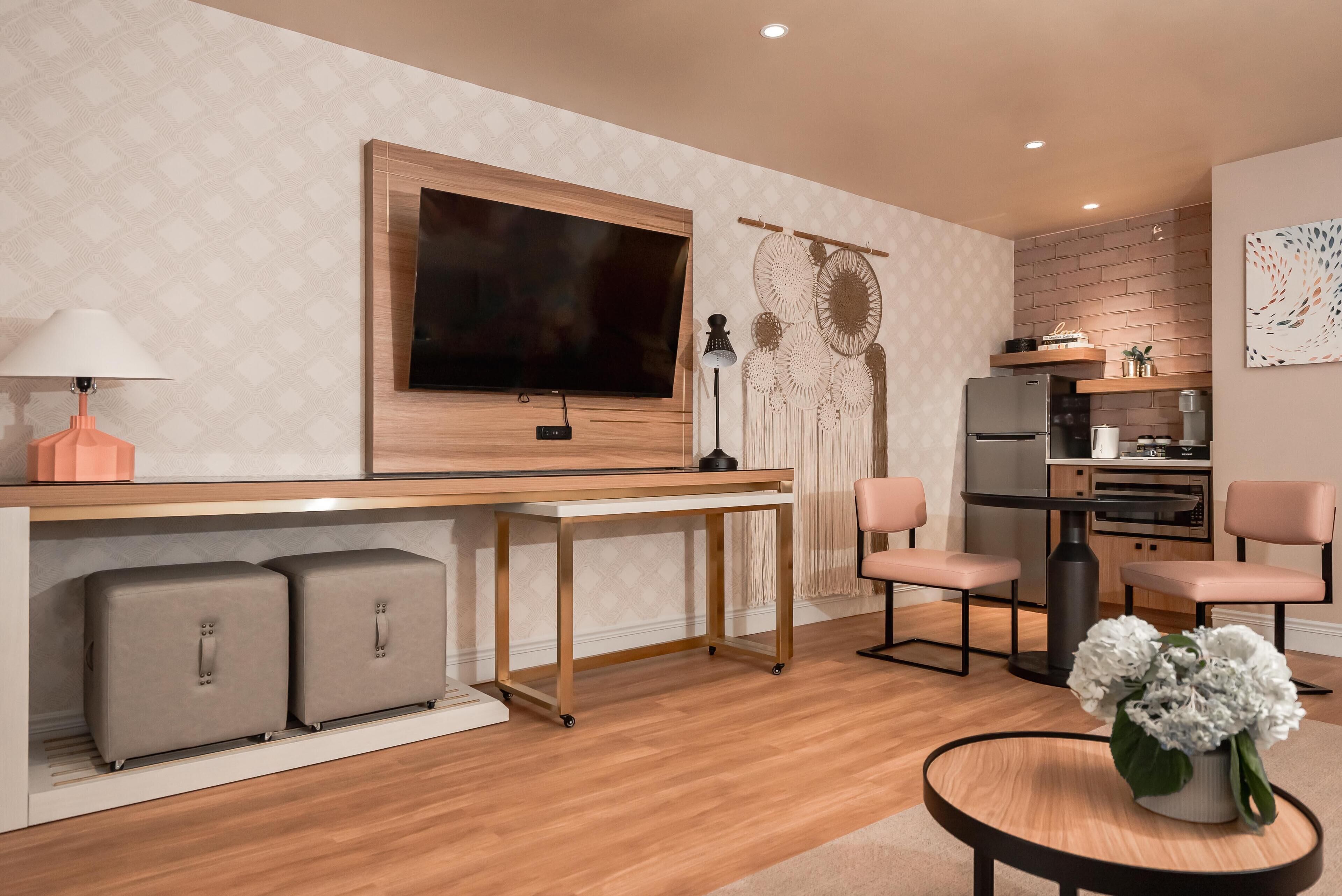 A modern living room with a wooden desk, a flat-screen TV, a lamp, and a dining area with chairs.