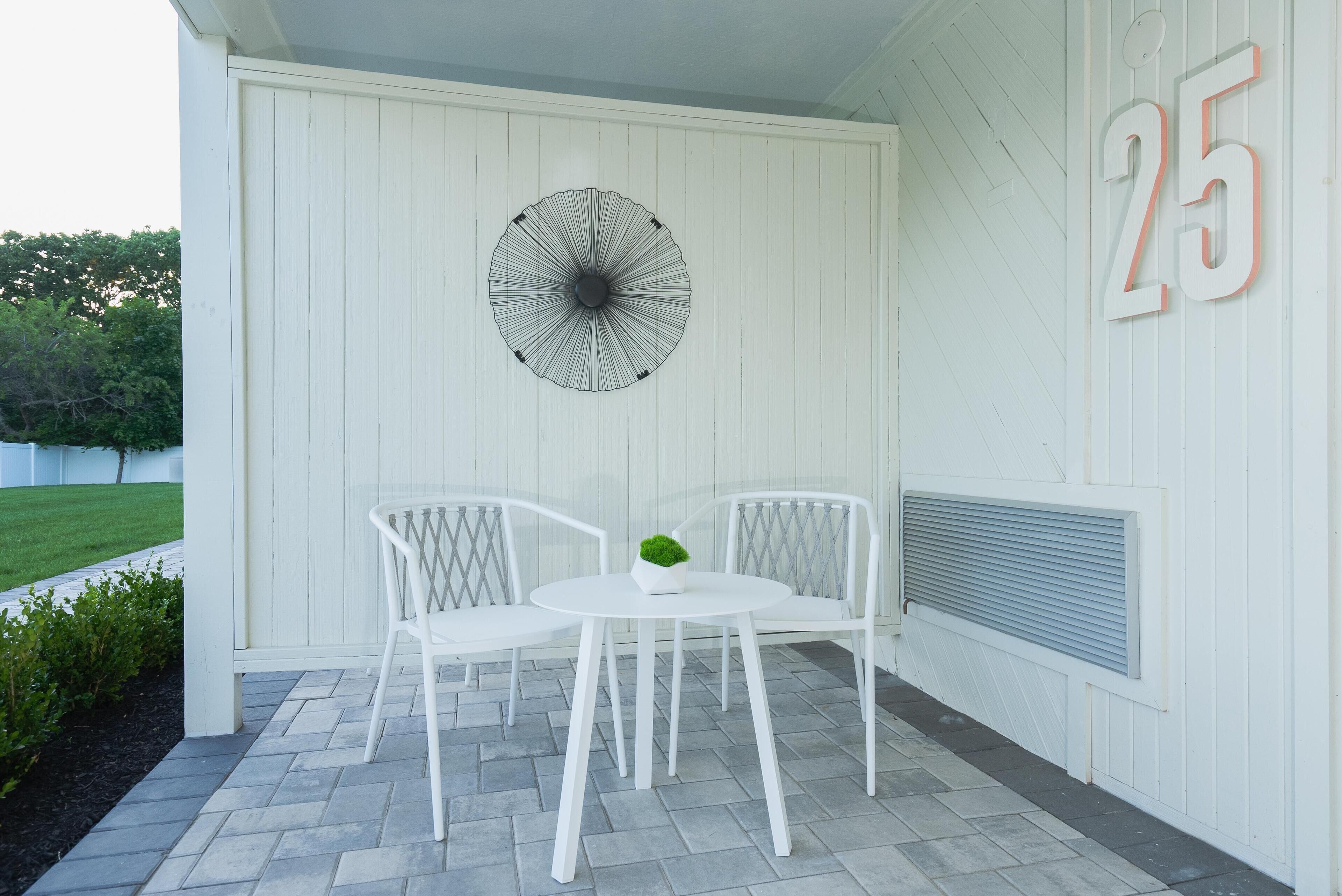 Een wit buiten terras met twee witte stoelen en een tafel, een decoratieve parasol, en een muur met het nummer 25.