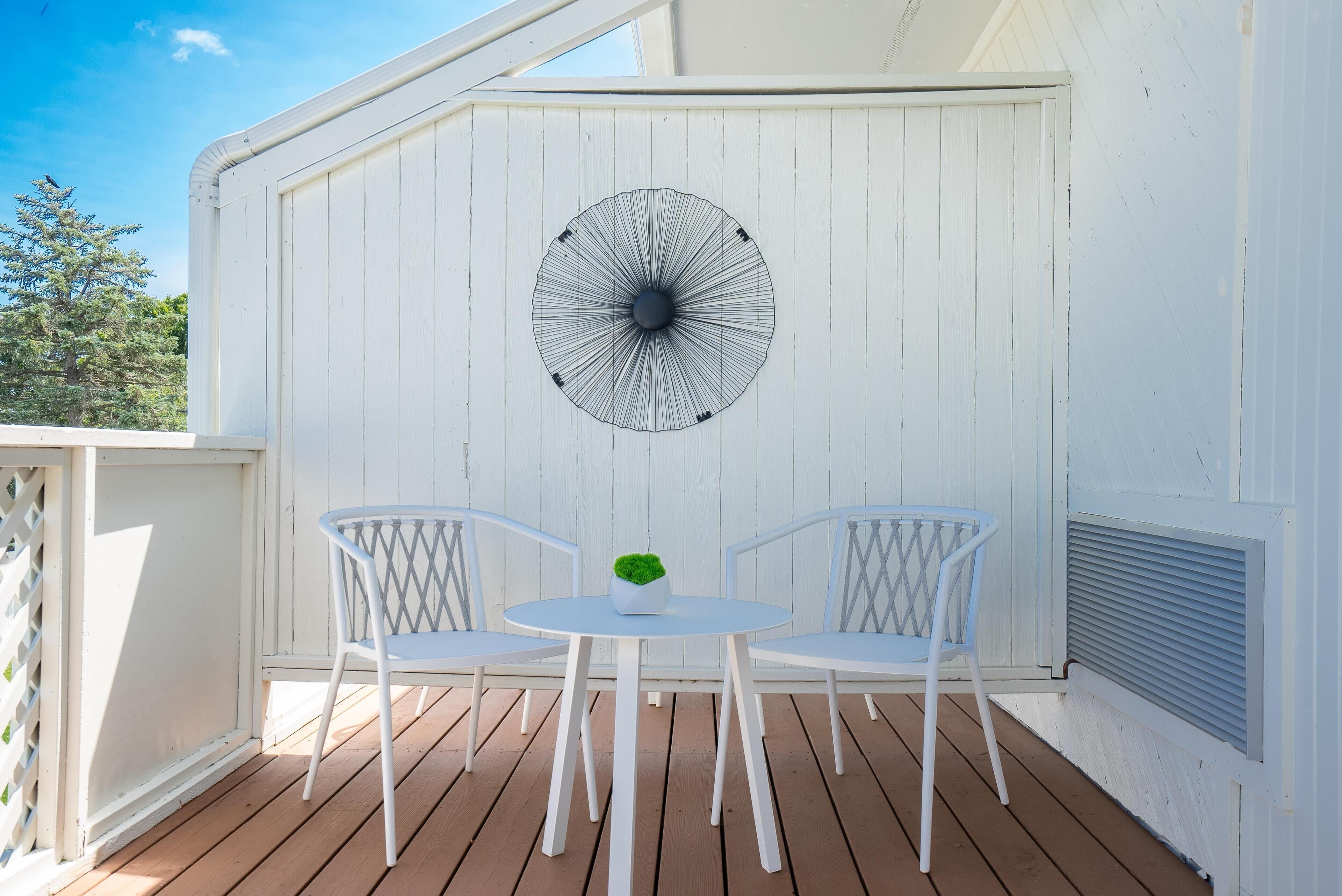 A white outdoor patio with two white chairs and a small table, featuring a decorative black and white fan on the wall.