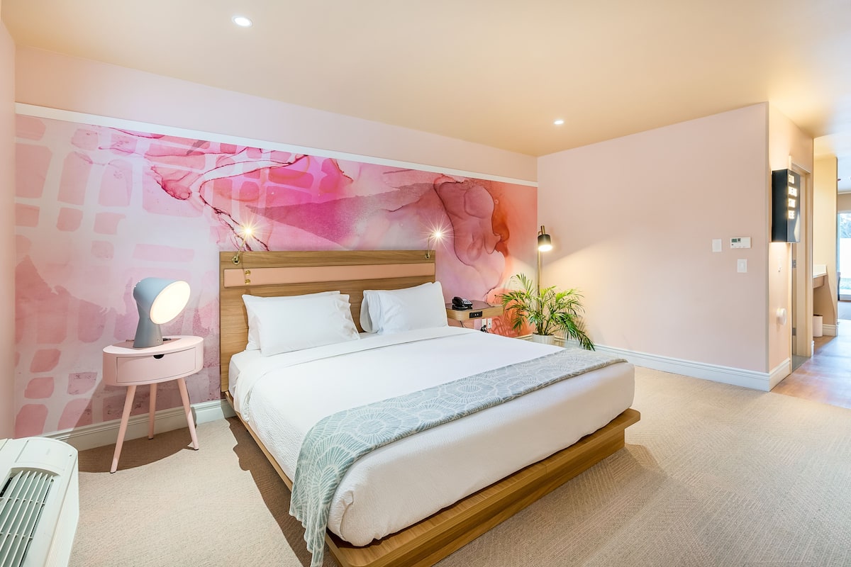 Une chambre avec un grand lit, une table de chevet avec une lampe, une fresque murale et une plante en pot.