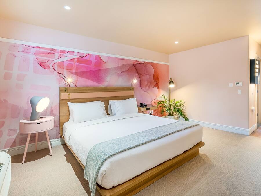 A bedroom with a large bed, a bedside table with a lamp, a wall mural, and a potted plant.