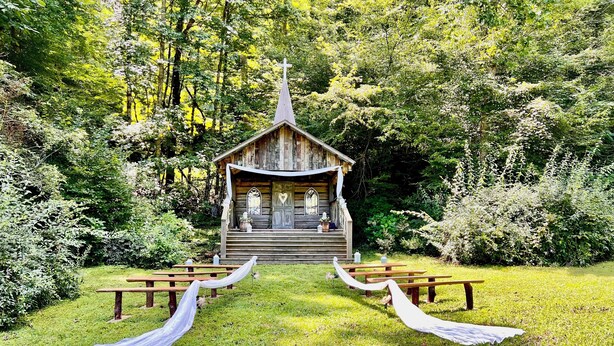 Una capilla de madera rústica con una cruz en la parte superior, rodeada de exuberante vegetación.