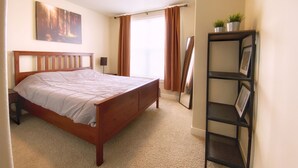 Une chambre avec un lit en bois, une table de chevet, une étagère avec des photos, une fenêtre avec des rideaux et un tableau accroché au mur.