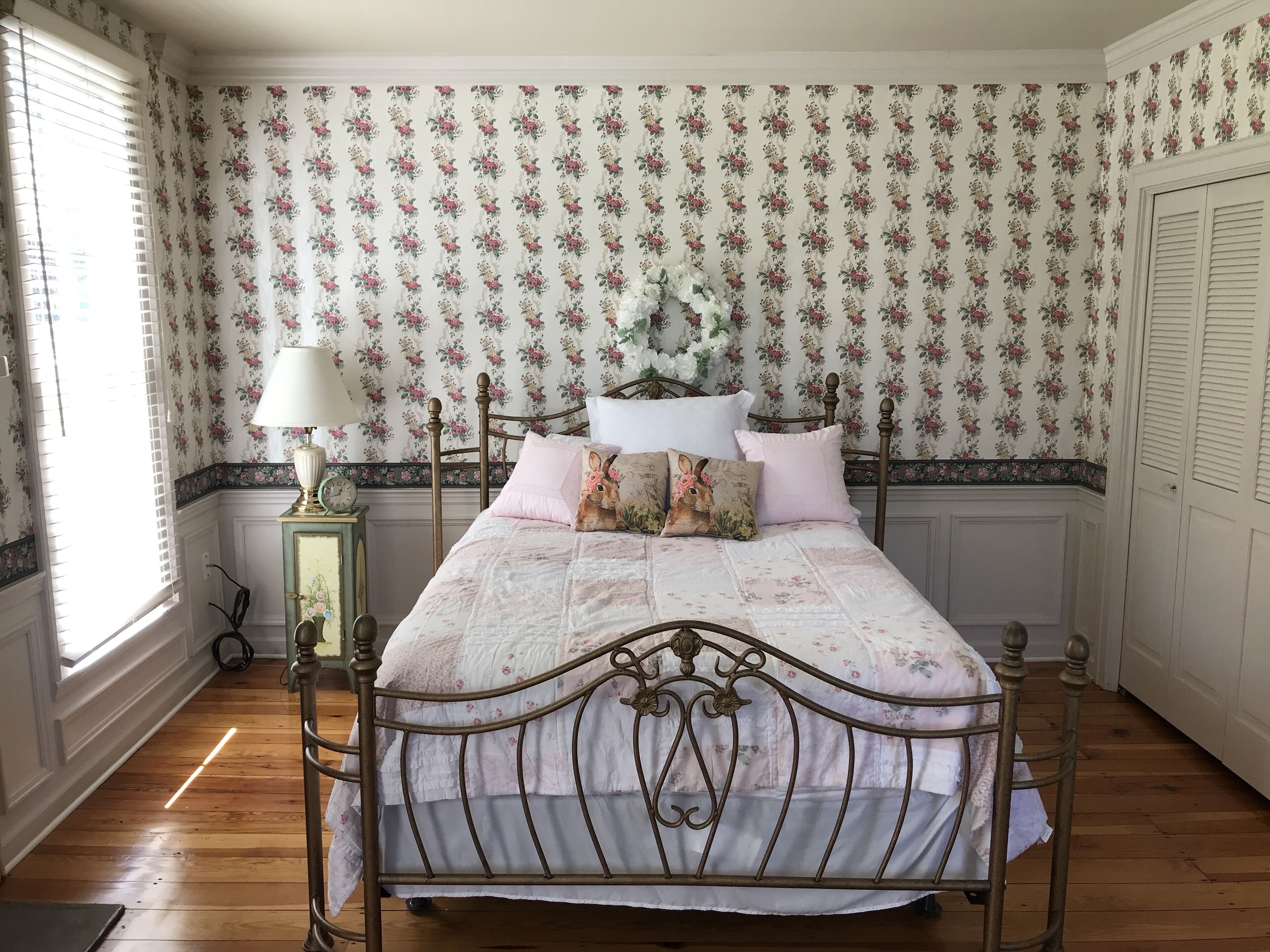 Un dormitorio con suelo de madera, papel tapiz floral, una cama con colcha blanca y rosa, y una mesita de noche con lámpara.