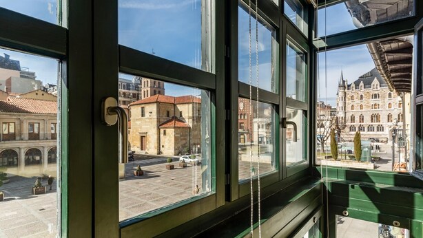 A view from inside a building with large windows overlooking a historic town square with buildings and a church. - Housing León- The Galery (León)