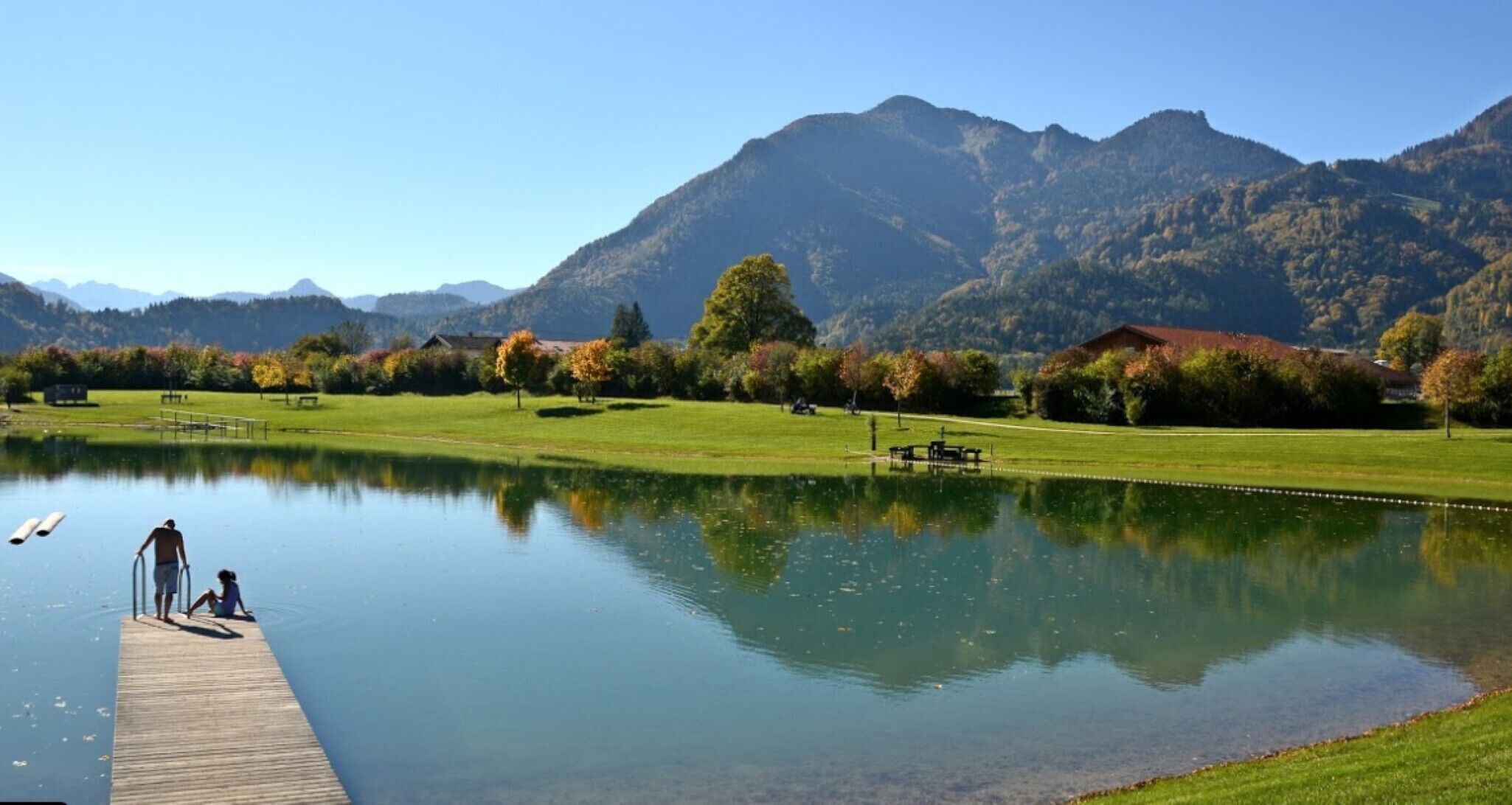 A calm lake with a wooden dock, surrounded by mountains and a grassy area with trees and a building.