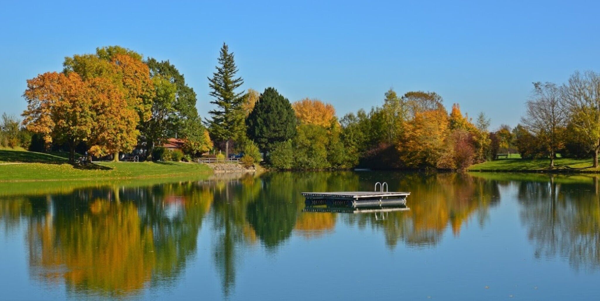 A calm lake with a dock, surrounded by trees with autumn foliage, reflecting the scenery.