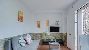 Ein Wohnzimmer mit grauem Sofa, einem kleinen Tisch mit Flasche und Gläsern, einem Flachbildfernseher an der Wand und einer Tür mit Vorhang.