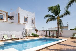 Zona de piscina con sillas de descanso, una palmera y un edificio blanco con ventanas.