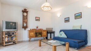 Ein Wohnzimmer mit blauem Sofa, einem Glastisch, einem Holzkasten mit Fernseher, einer Uhr und gerahmten Bildern an der Wand.