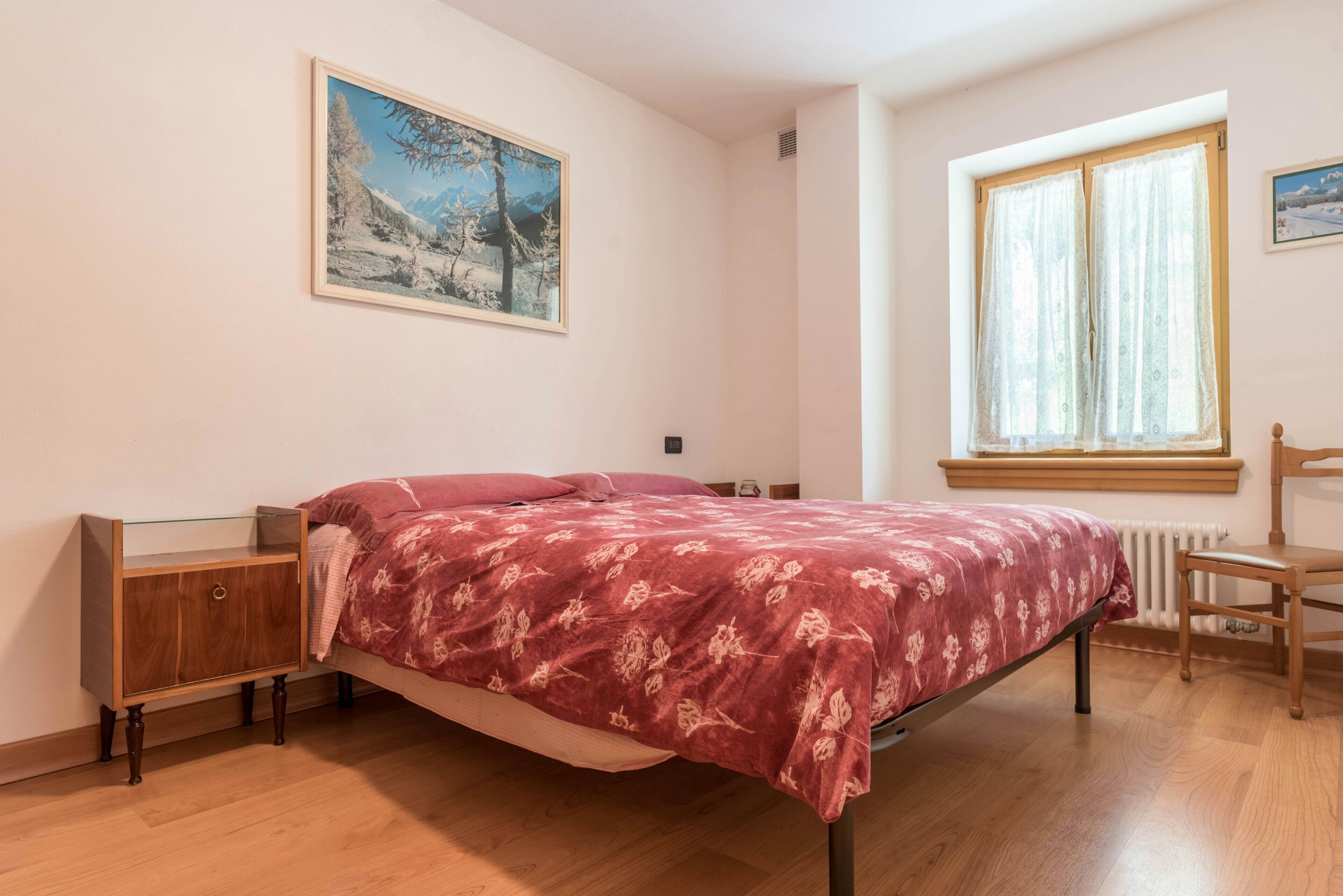 A bedroom with a bed, a nightstand, a window with curtains, a radiator, and a painting on the wall.