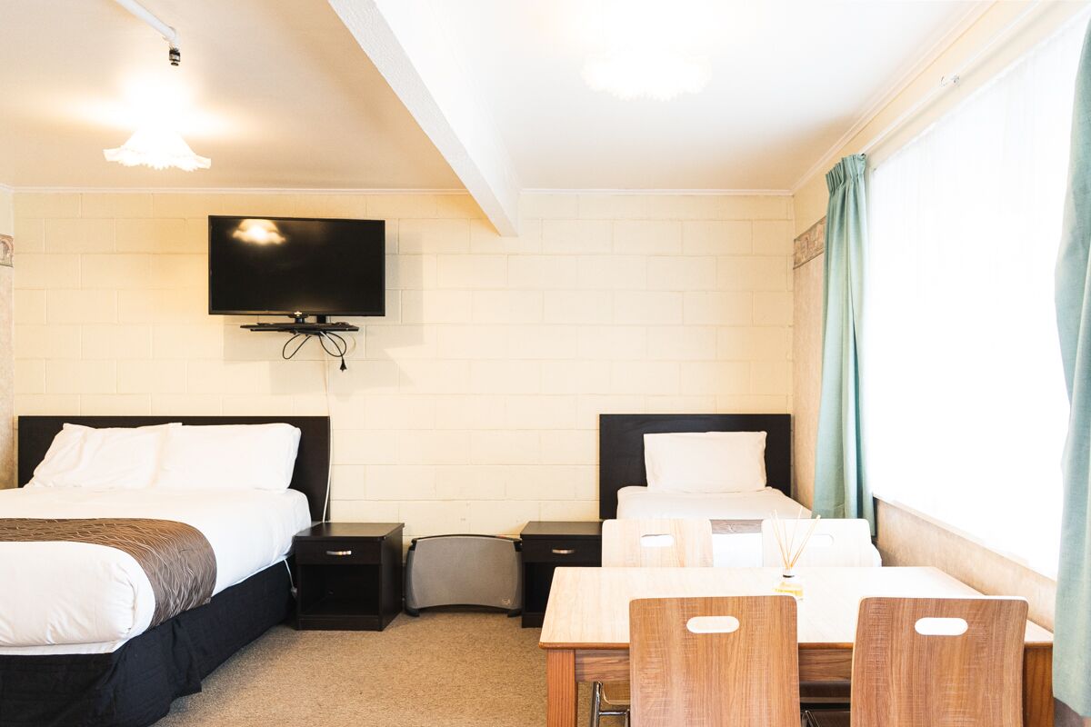 A hotel room with two beds, a TV mounted on the wall, a desk with chairs, and a window with curtains.