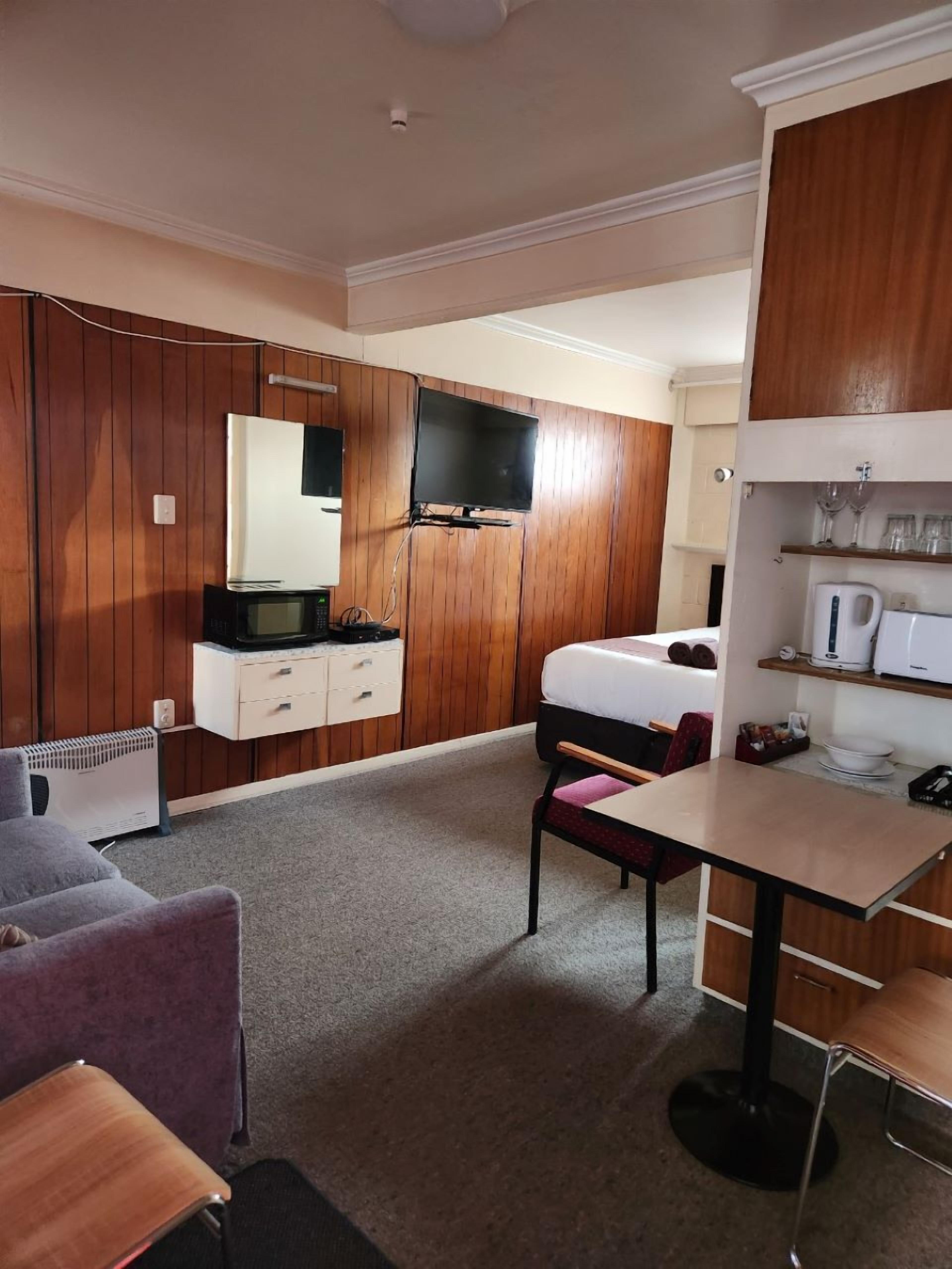 Habitación de hotel con armario de madera, televisor de pantalla plana, cama, escritorio con silla y una pequeña zona de kitchenette.