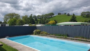 Une piscine rectangulaire, entourée d’une clôture sombre, se détache sur un paysage de collines verdoyantes et d’arbres, sous un ciel nuageux.