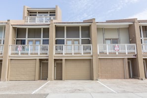 A multi-story residential building with three garage doors, white railings on balconies, and a clear sky.