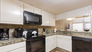 Una cocina con gabinetes blancos, microondas negro, estufa, cafetera y lavavajillas.