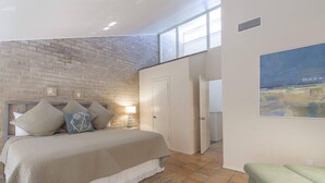 Un dormitorio con pared de ladrillo, cama con almohadas, mesita de noche, un cuadro de playa y una silla verde.