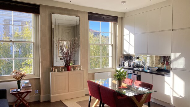 Une cuisine moderne avec des meubles blancs, une table à manger en verre et des chaises rouges. Une grande fenêtre offre une vue sur les arbres et un bâtiment à l’extérieur.