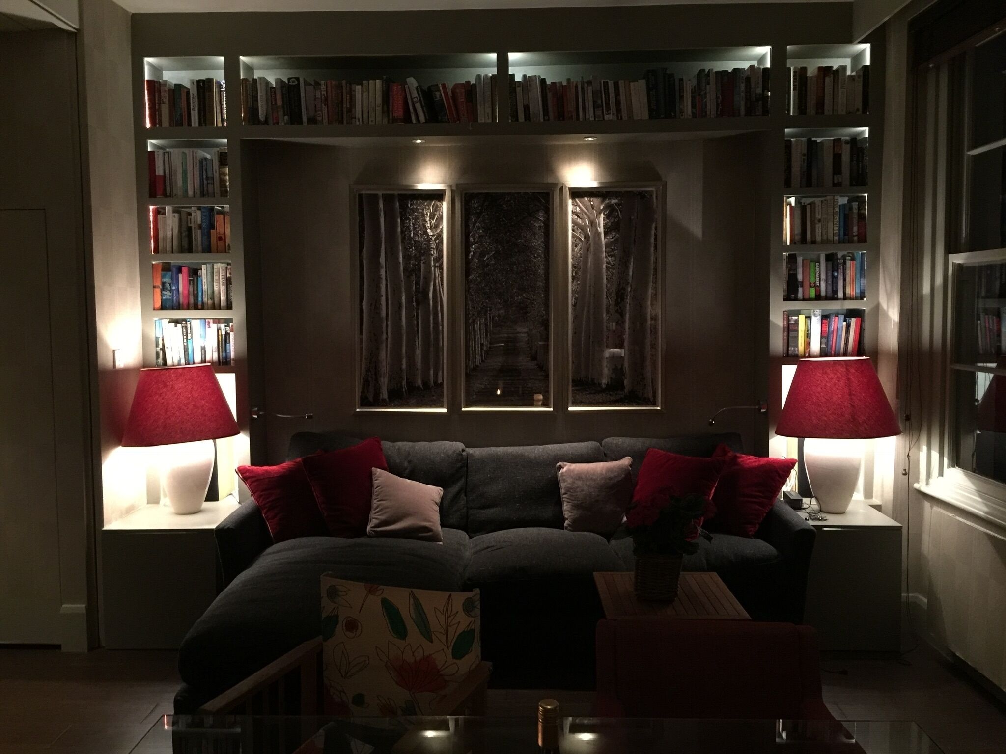 Een woonkamer met een donkergrijze bank, rode kussens, een glazen salontafel en een boekenkast vol boeken. Er zijn twee ramen met gordijnen, en een rode lamp aan elke kant van de bank.