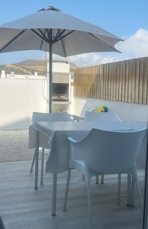 Une terrasse extérieure avec une table et des chaises sous un grand parasol, un barbecue et une clôture en bois.