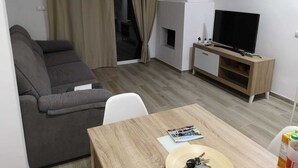 Een moderne woonkamer met een grijze bank, een houten salontafel, een televisie op een houten dressoir en een glazen schuifdeur met gordijnen.