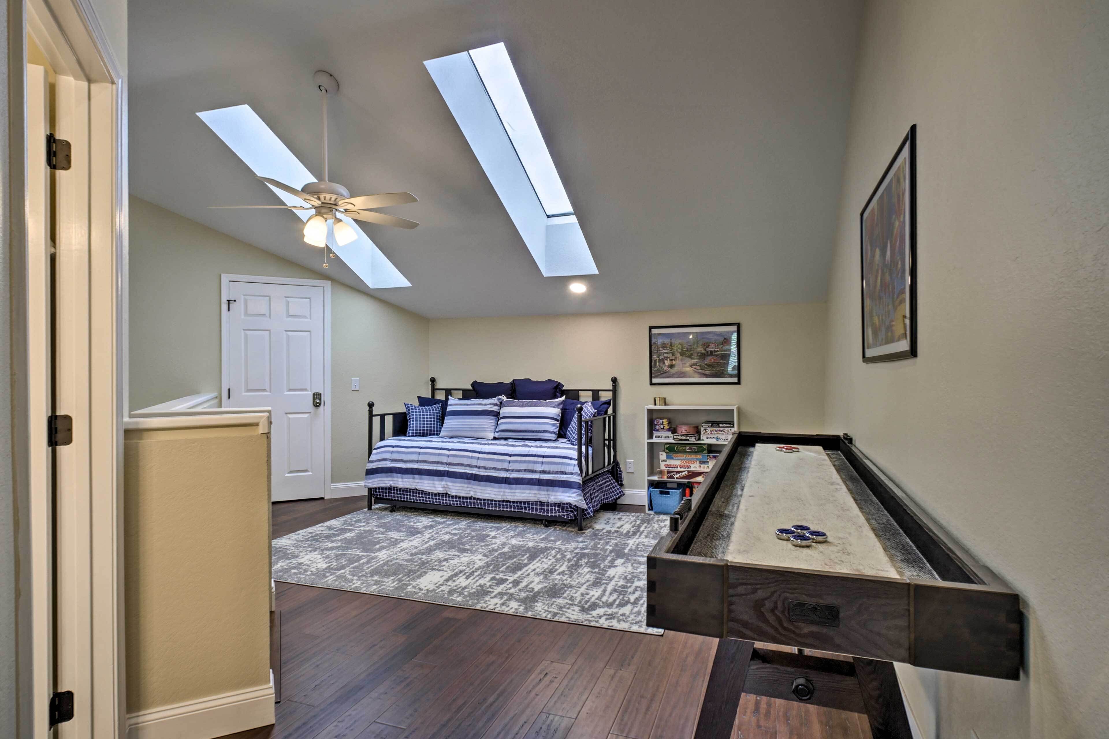 A bedroom with a bed, a ceiling fan, skylights, a door, a bookshelf, and a pool table.