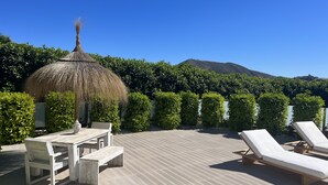 A wooden deck with lounge chairs and a table, surrounded by neatly trimmed hedges and a thatched-roof structure. - A truly exceptional villa (Xàbia)