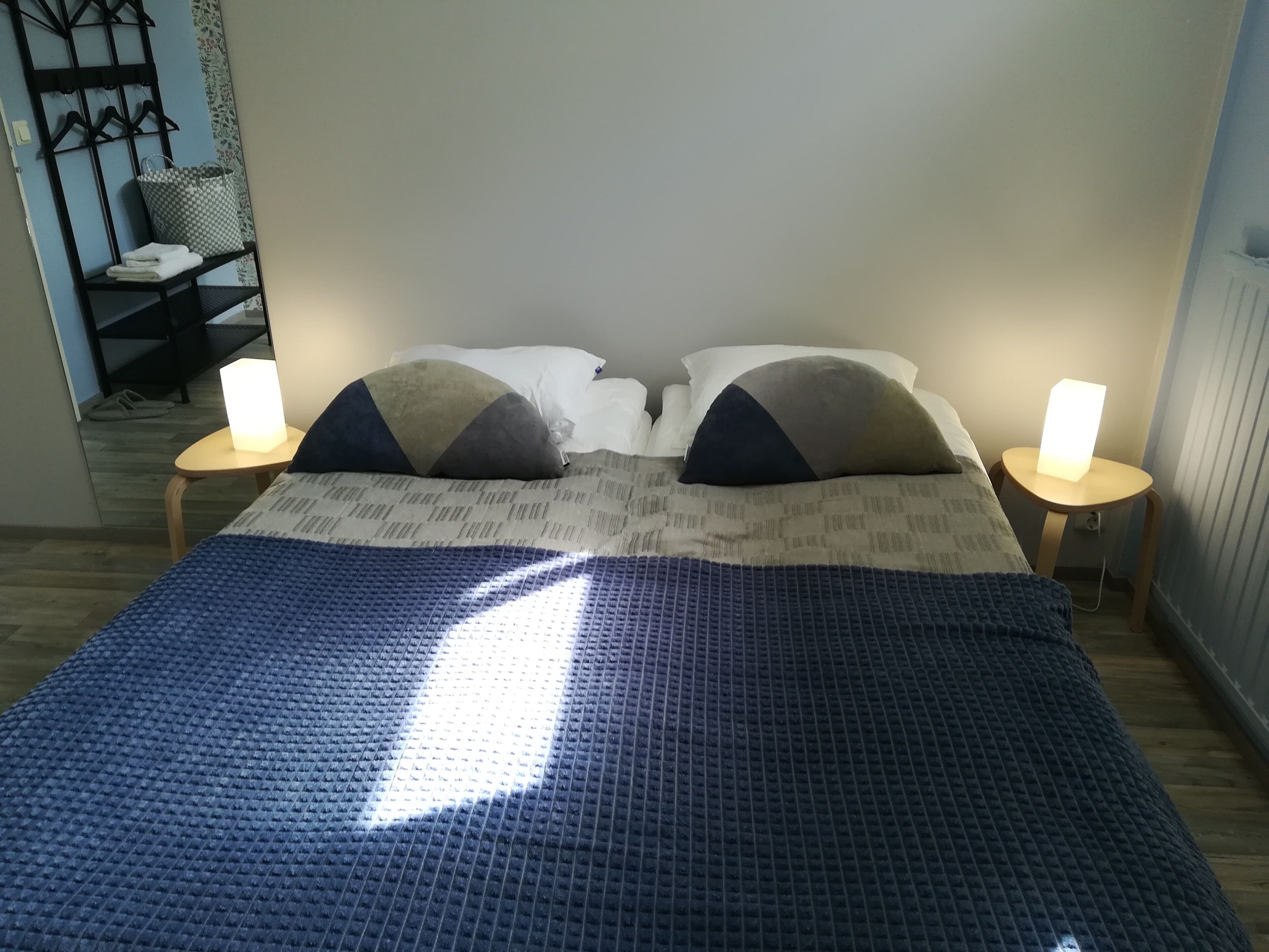 A bed with a blue textured cover, two bedside tables with lamps, and a wall-mounted rack with hangers.