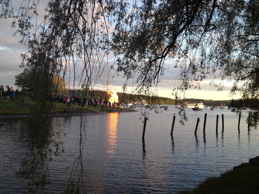 A lakeside view with a crowd gathered near a fire, boats on the water, and trees in the foreground.