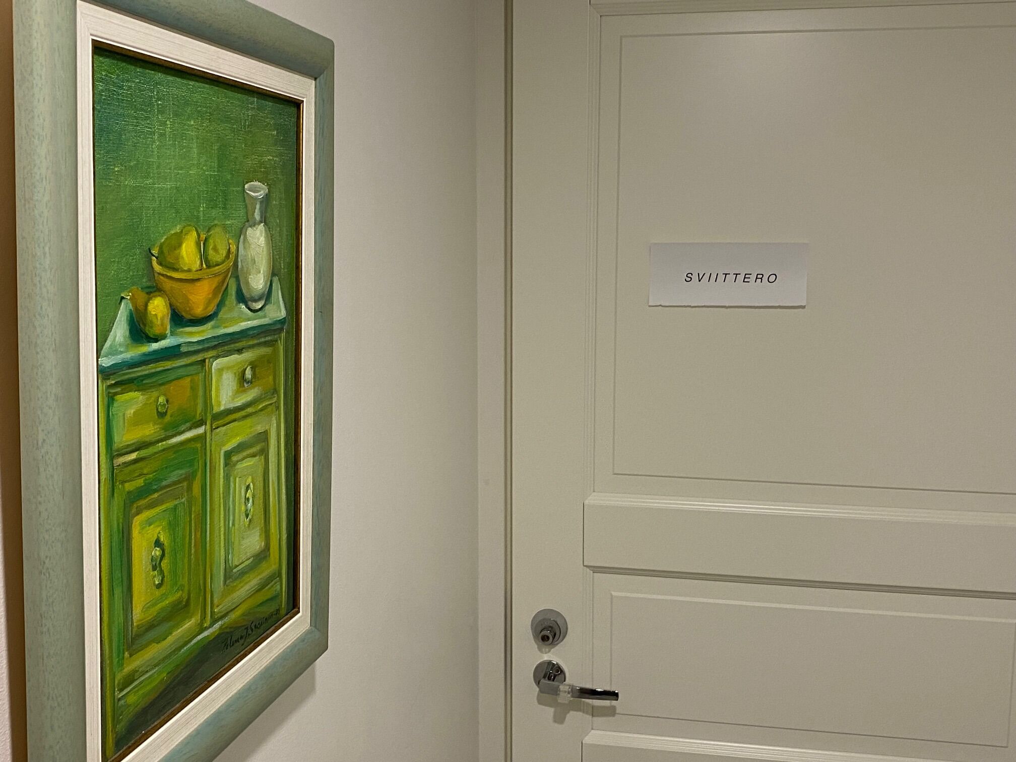 Une porte sur laquelle est apposée une enseigne indiquant « SVITTERO », ainsi qu’un tableau représentant un buffet vert avec un saladier et des citrons.