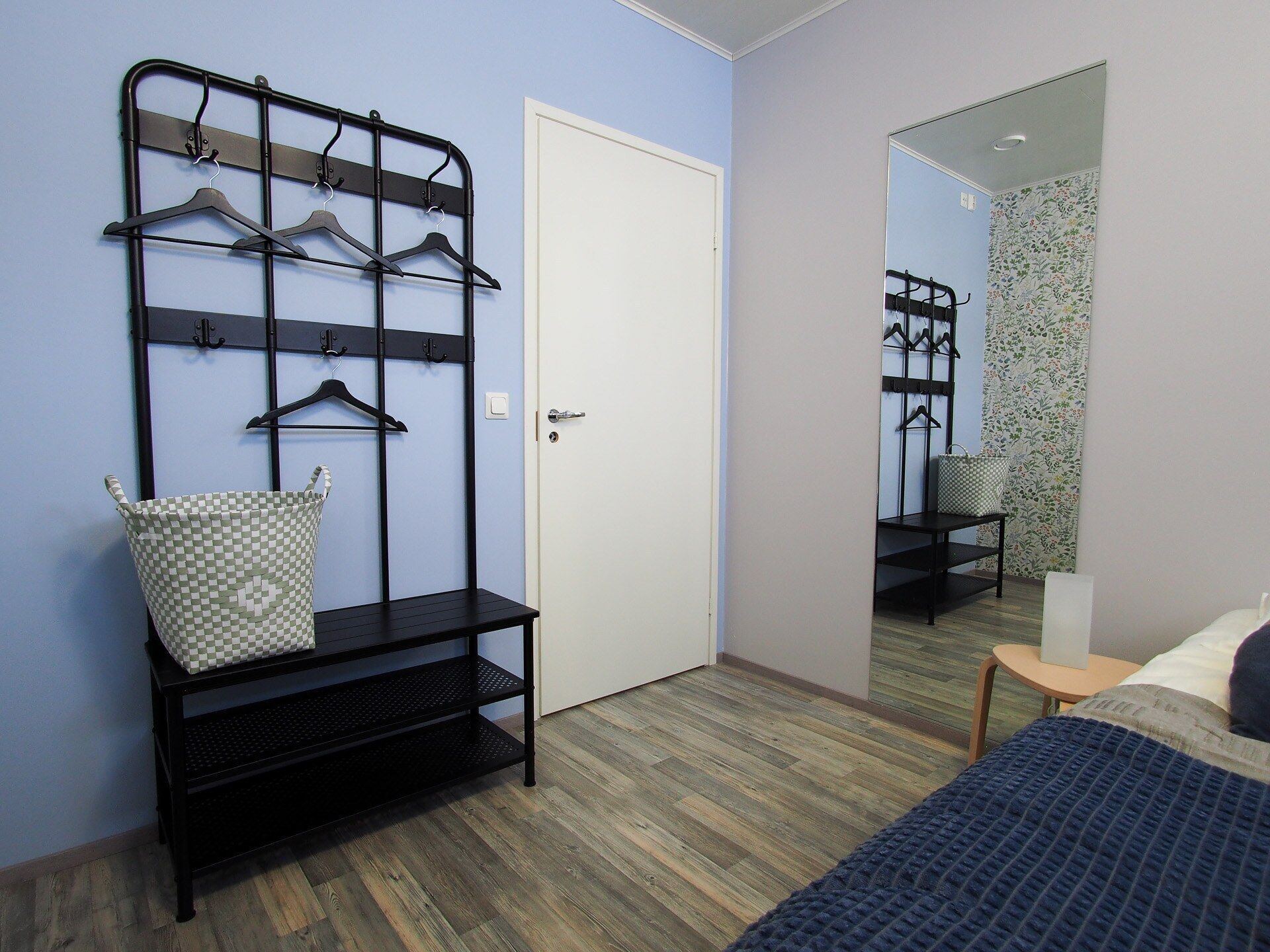 A bedroom with a bed, a mirror, a wardrobe with a hanging rack, and a small table with a basket.
