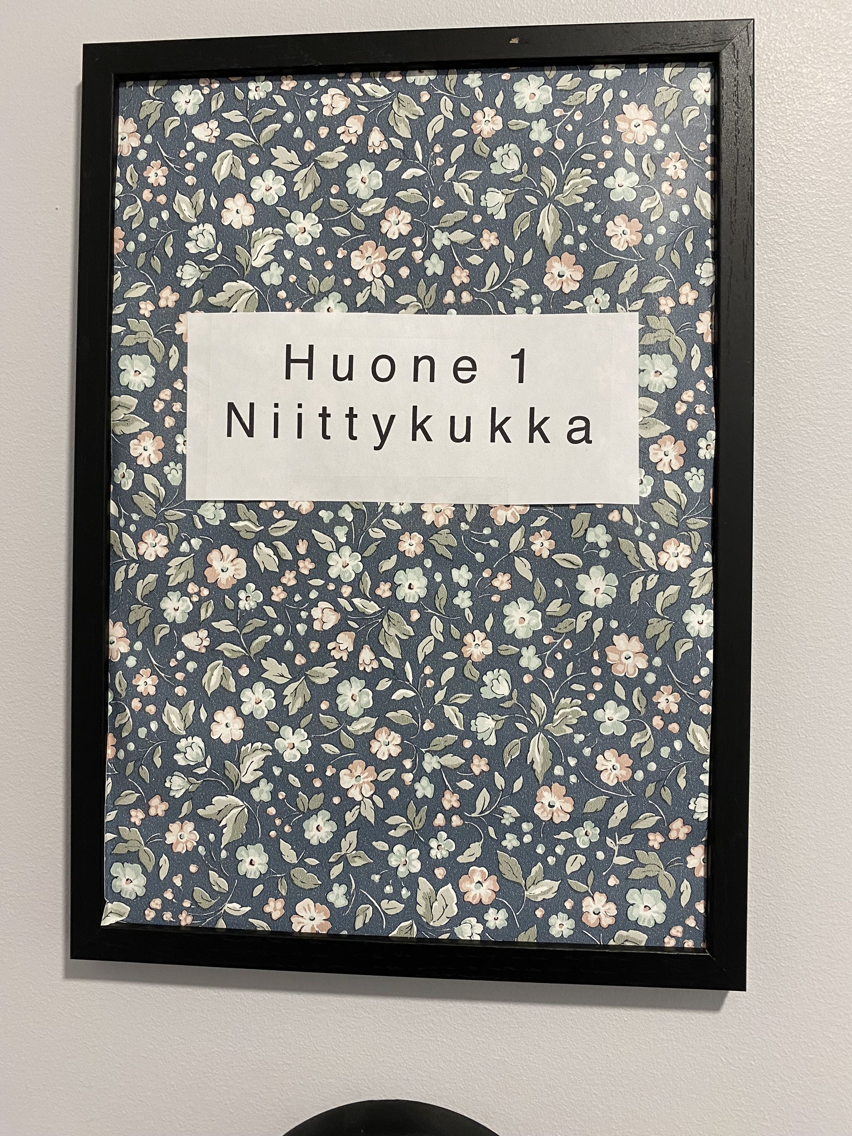 A framed artwork with a floral pattern and a label that reads "Huone 1 Nittykukka".