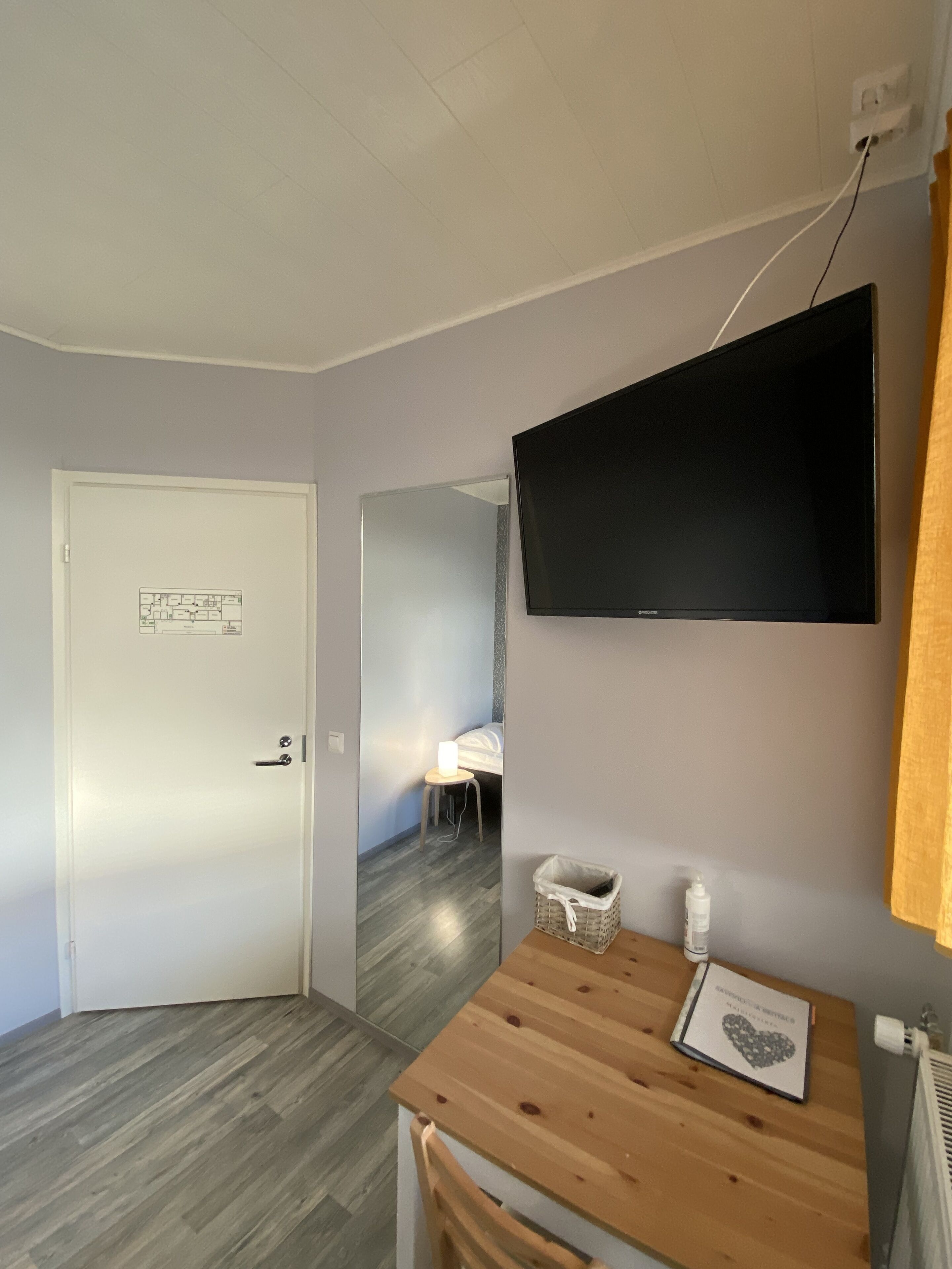 Una habitación con una mesa de madera, un televisor de pantalla plana montado en la pared, una puerta blanca y un espejo que refleja una cama y una mesita de noche.