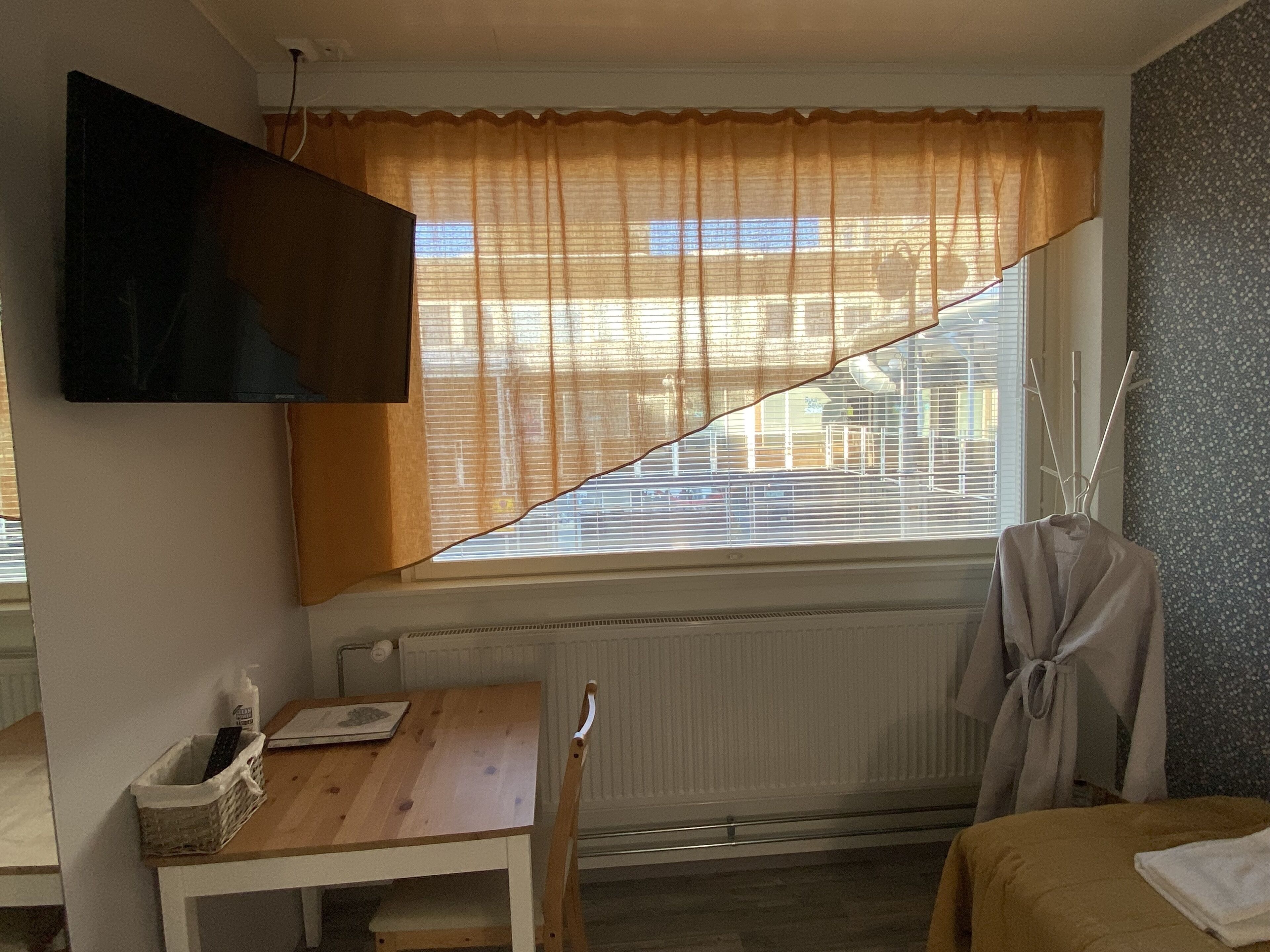 A room with a wooden table, a chair, a bed, a TV mounted on the wall, and a window with curtains.