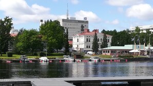 A waterfront area with boats docked, a large building with a red roof, and a green-roofed structure with flags. - Savonlinna Rentals (Savonlinna)