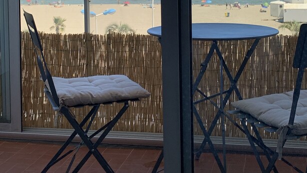 En balkong med två stolar och ett bord, med utsikt över en strand med parasoller och folk.