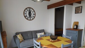A cozy living room with a gray sofa, a wooden dining table with chairs, a wall clock, and a small kitchenette area.