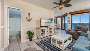 A spacious living room with a television, a ceiling fan, a large window with a view of the ocean, and a balcony with a view of the ocean.