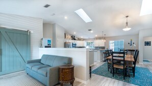 A spacious living room with a sofa, a dining area with a table and chairs, a kitchen with white cabinets, and a large window. - Best Family Vacation at the Beach or the Pool. (Tarpon Springs)