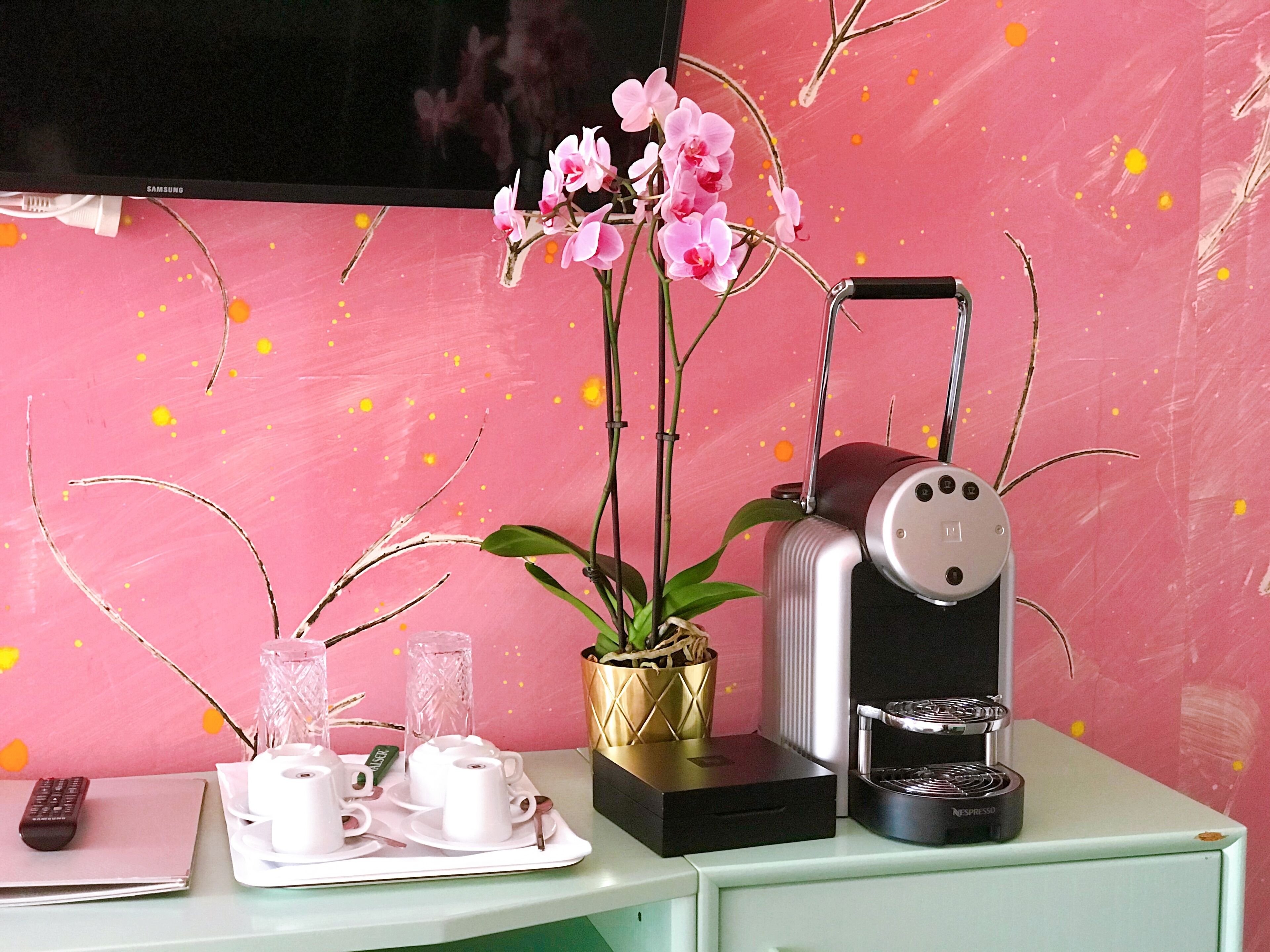 Ein Schreibtisch mit Kaffeemaschine, Fernbedienung, Tablett mit Tassen und einer Vase mit Blumen.