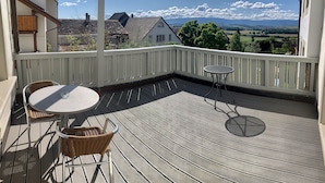 A wooden deck with a round table and two wicker chairs, overlooking a scenic landscape with houses and mountains. - Swiss Hotel La Couronne (Avenches)