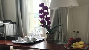 A hotel room with a dining table set with white cups and saucers, a tray with a fruit bowl containing bananas and grapes, a potted orchid, and a lamp. - Swiss Hotel La Couronne (Avenches)