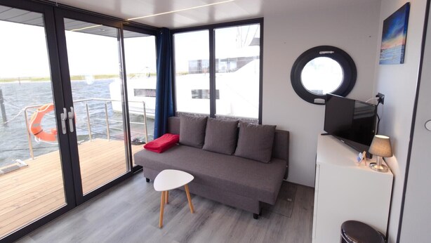 A modern room with a grey sofa, a small white table, and a TV on a stand. - Houseboat Fjord Aquila with roof terrace in Wendtorf (Wendtorf)