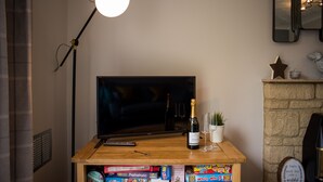Un mueble de televisión de madera con un televisor de pantalla plana, una botella de champán y un control remoto. Debajo del mueble, hay juegos de mesa y una pequeña planta en maceta.