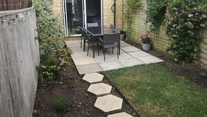 Un patio con mesa y sillas, una puerta corrediza de vidrio y un jardín con diversas plantas.