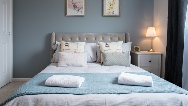 Una cama bien hecha con una colcha azul claro y dos toallas blancas, un cabecero tapizado en gris y dos cuadros en la pared.